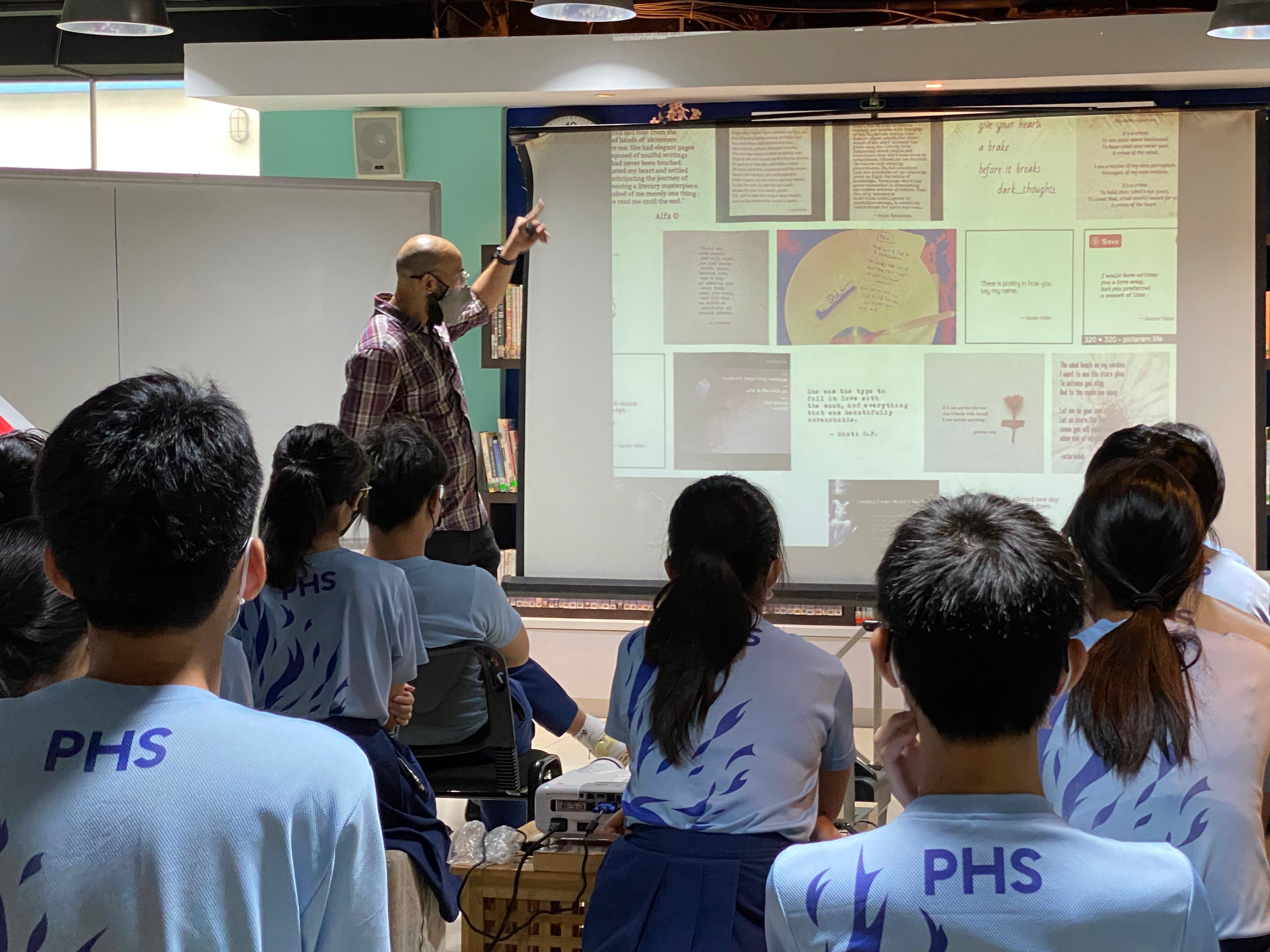 A teacher points at a projection screen showing poetry quotes while PHS uniformed students watch from behind.